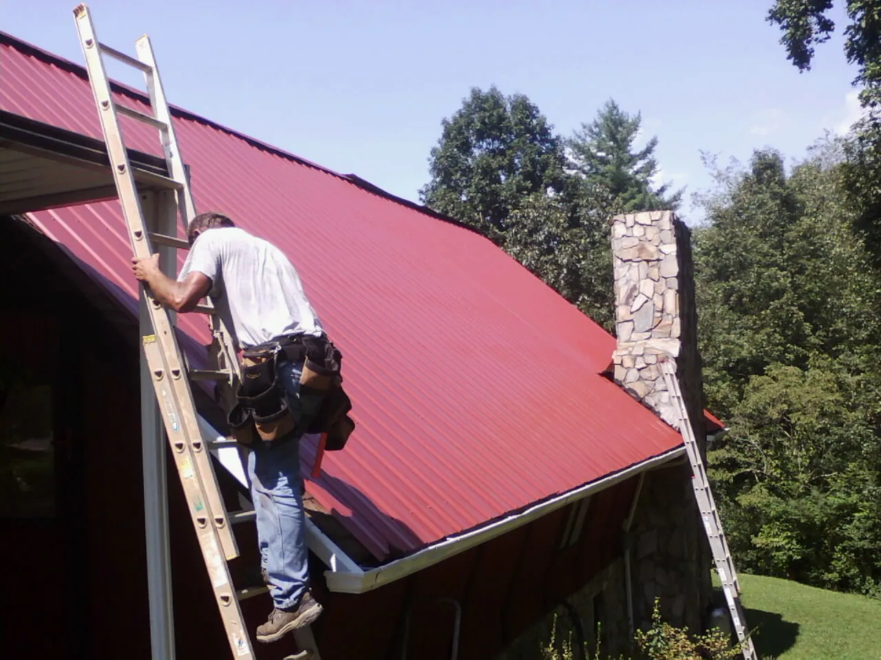 Roofer on a ladder beside a red metal roof and stone chimney — Soffit & Fascia Repair by Roofing Brothers in Carbon